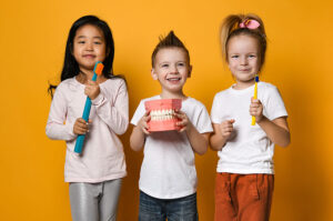 Three children holding toothbrushes and a dental model, smiling against a bright yellow background, emphasizing the importance of pediatric dental health and early dental visits.