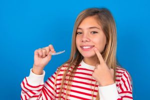 Young girl in striped sweater holding clear aligner and pointing to her smile, representing cosmetic dental solutions for developing teeth.