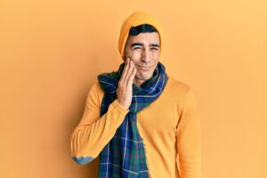 Man in orange sweater and yellow beanie holding his jaw, showing signs of dental discomfort against a solid orange background.