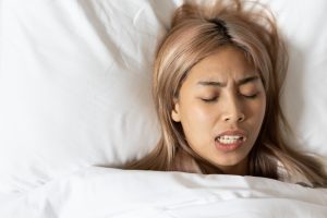 Woman with long, light-colored hair grimacing in bed, indicating discomfort possibly related to teeth grinding or bruxism, relevant to dental care and protection for veneers.
